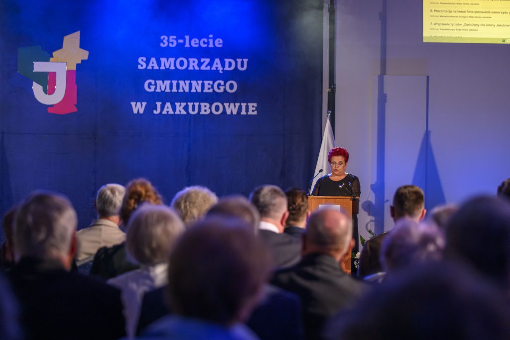 Na scenie kobieta przemawia przed publicznością. Tło jest niebieskie z napisem o 35-leciu samorządu. Na ekranie po prawej prezentacja.