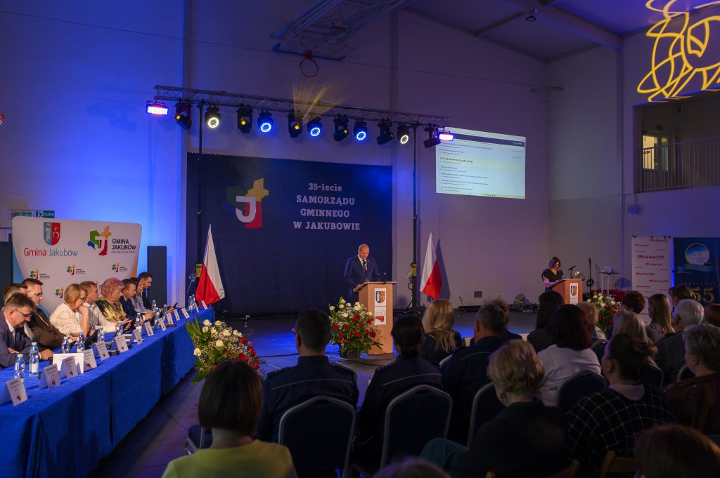 Spotkanie z okazji 35-lecia samorządu w Jakubowie. Scena z przemawiającym mężczyzną, niebiesko-czerwone oświetlenie. W tle flagi i projektor.