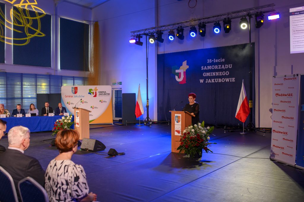 Sala konferencyjna z niebieskim oświetleniem, przemawiająca osoba przy mównicy, na ścianie napis 35-lecie Samorządu Gminnego w Jakubowie.