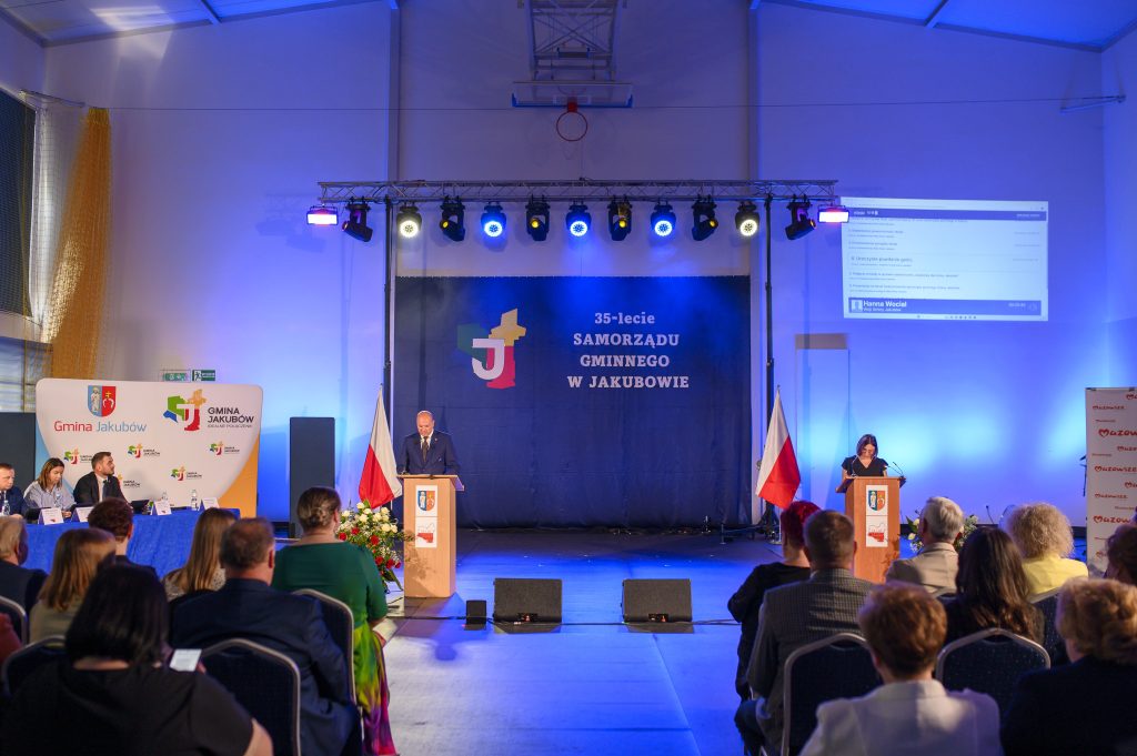 Na zdjęciu widać salę z niebieskim oświetleniem, podium i flagi Polski. Tło jest ciemnoniebieskie z napisami. Ludzie siedzą na krzesłach, słuchając przemówienia.