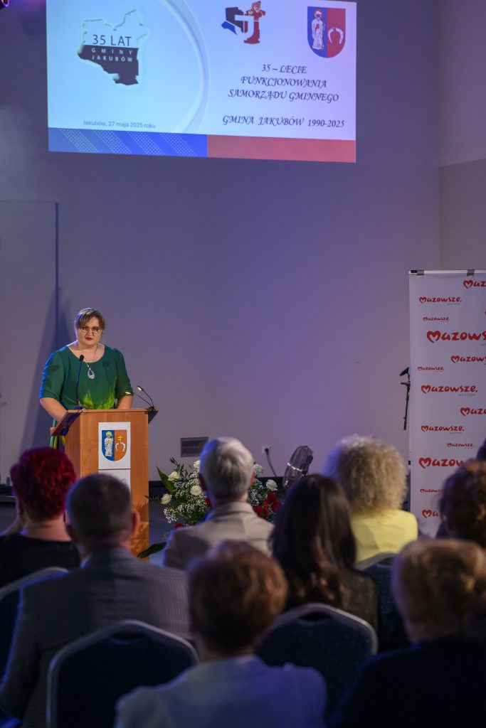 Osoba przy mównicy w zielonej sukience przemawia. Na ekranie z tyłu logo i napisy o 35-leciu gminy Jakubów. W tle banner z napisem Mazowsze.