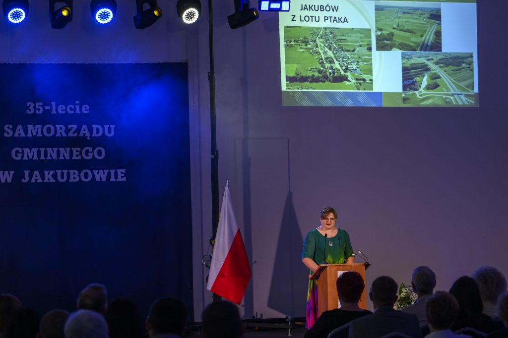 Na scenie kobieta w zielonej sukni przemawia przy mównicy. Tło niebieskie z białym napisem o rocznicy samorządu gminnego. Obok polska flaga, nad nią slajd z widokiem z lotu ptaka.
