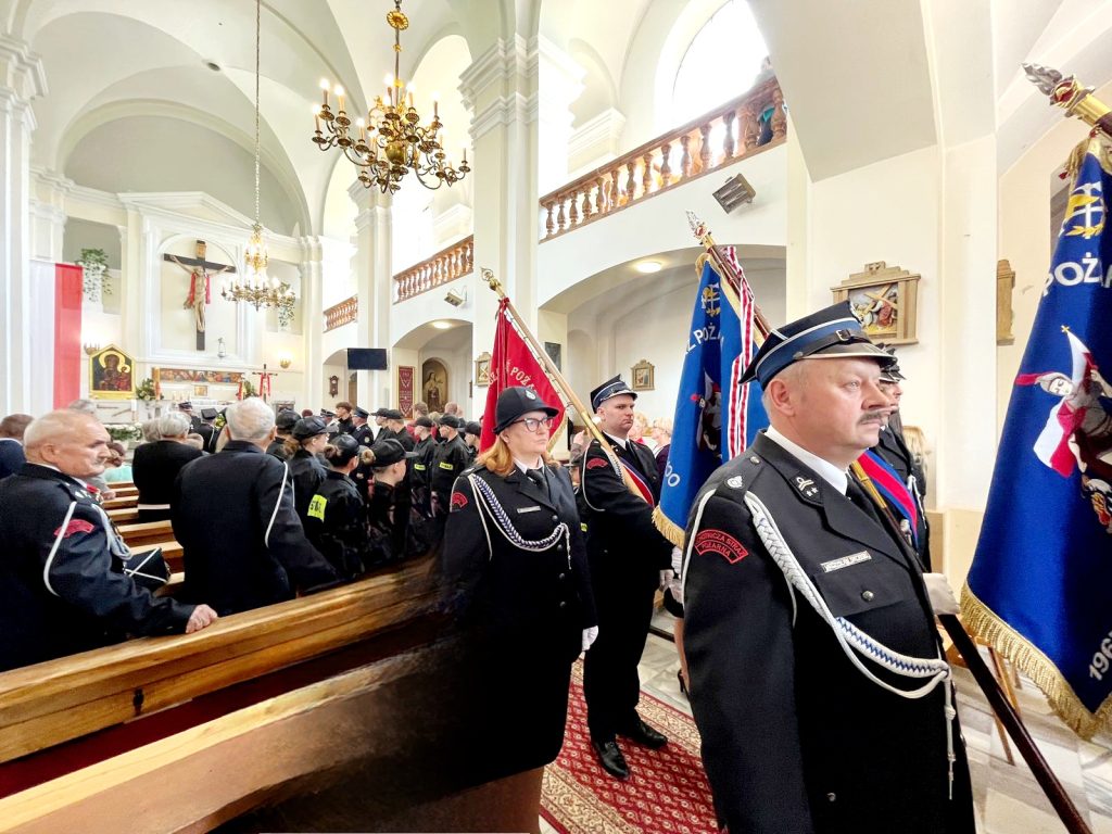 Grupa strażaków w mundurach stoi w jasnym kościele z białymi ścianami. Trzymają kolorowe sztandary. W tle widać ołtarz i żyrandole.