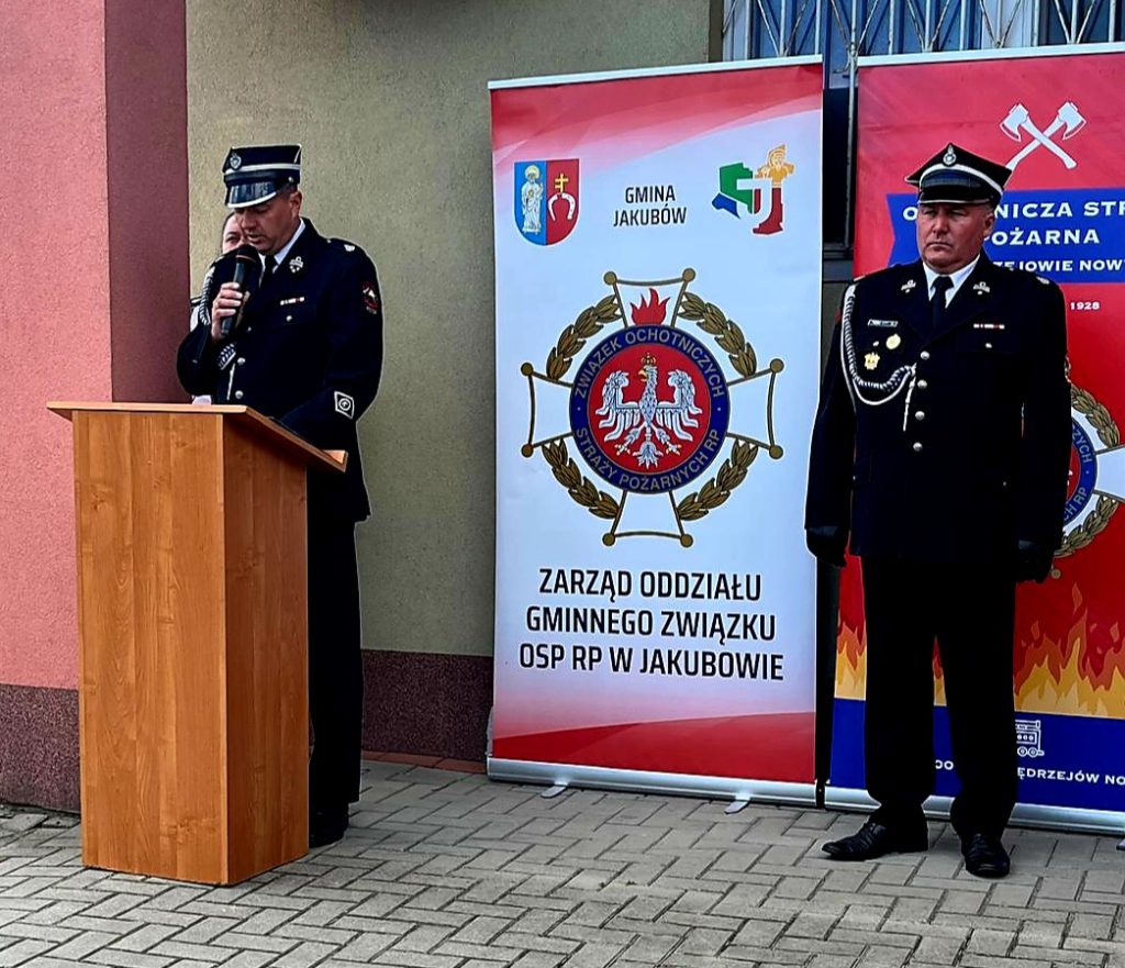 Na zdjęciu są dwaj strażacy w mundurach przy sztandarach OSP. Jeden przemawia przy drewnianej mównicy. Tło jest w kolorze czerwonym i szarym.