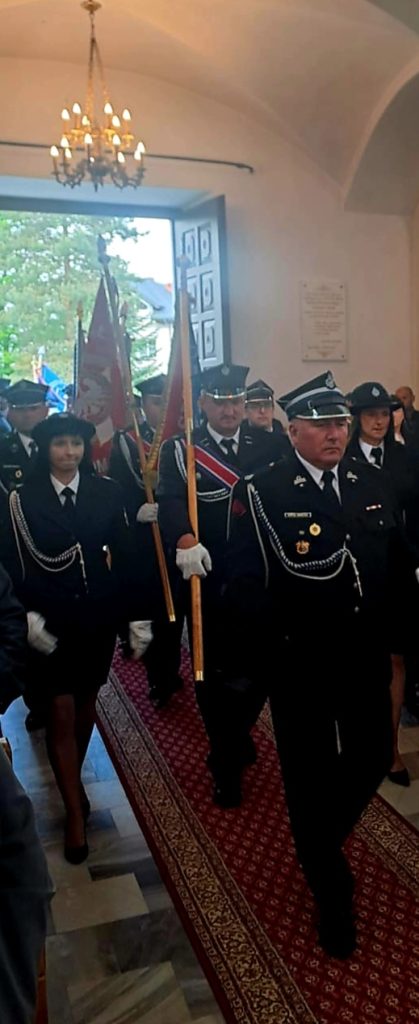 Grupa strażaków w galowych mundurach wchodzi do wnętrza budynku. Niosą flagi, idąc po czerwonym dywanie. W tle widać otwarte drzwi i chandler.
