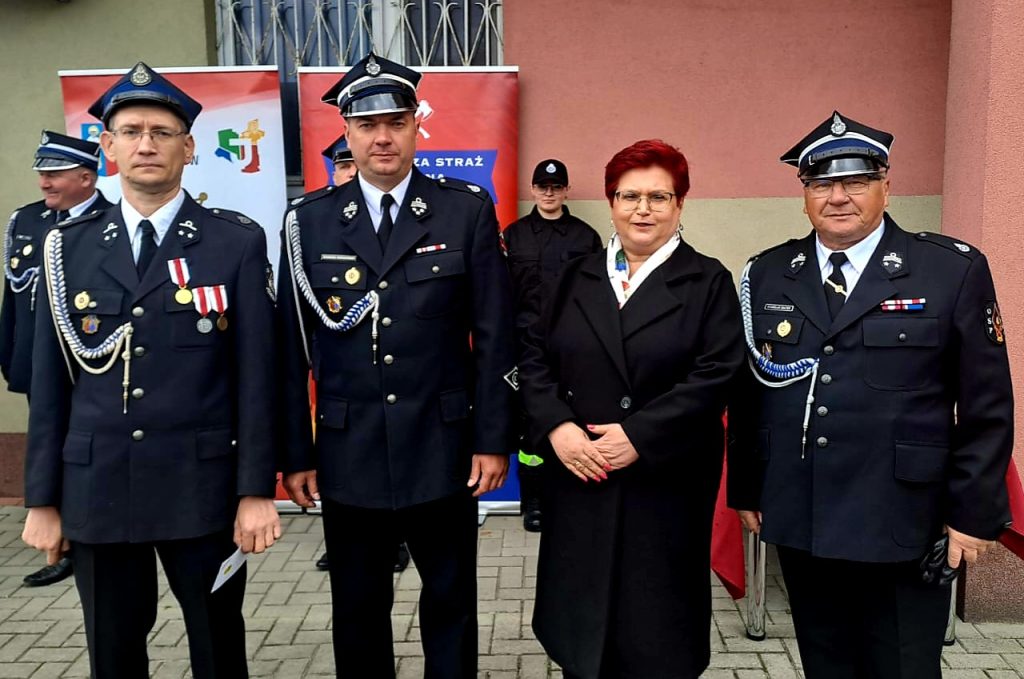 Na zdjęciu grupa ludzi w mundurach strażackich na tle budynku o różowej ścianie. Mają ciemne mundury z medalami i czapki. W tle są czerwone banery oraz częściowo zasłonięte postacie.