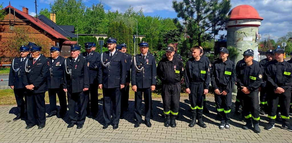 Grupa strażaków w mundurach stoi na brukowanej powierzchni. W tle zielone drzewa i czerwony dach budynku. Pogoda słoneczna, niebo częściowo zachmurzone.