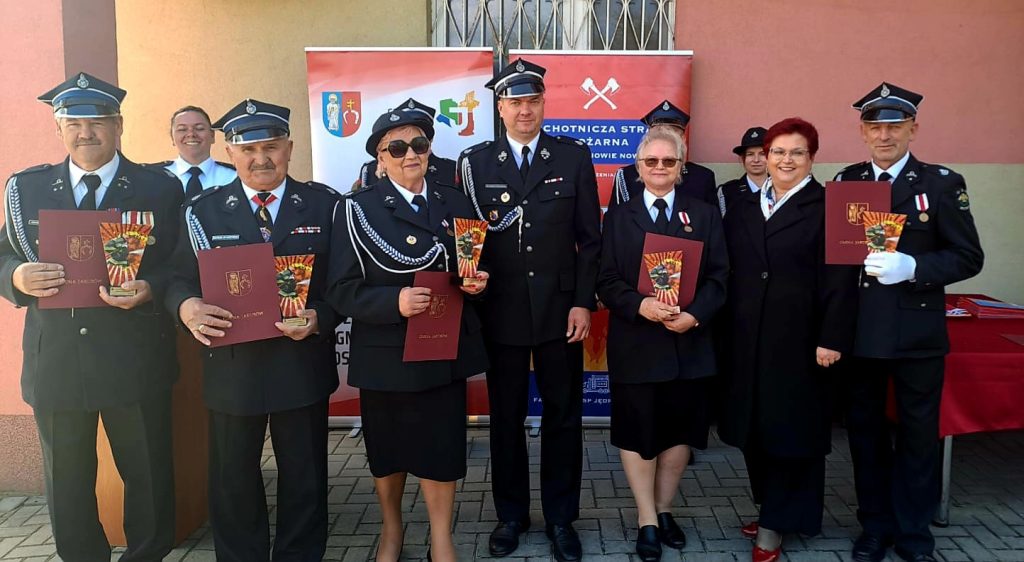 Grupa strażaków w mundurach z odznaczeniami i dyplomami. Tło stanowi kolorowy baner na pomarańczowej ścianie. Uroczystość odbywa się na zewnątrz.