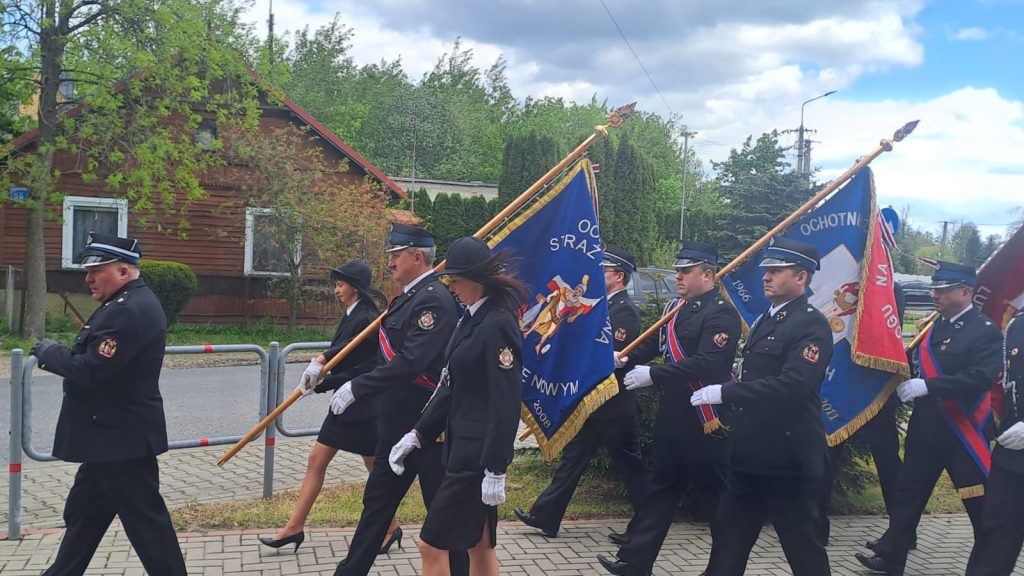 Grupa osób ubranych w mundury niesie kolorowe sztandary. Tło: zielone drzewa, brązowy dom. Chodnik i niebo z chmurami.