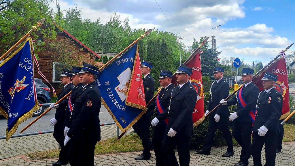 Grupa strażaków w ciemnych mundurach maszeruje, trzymając kolorowe flagi. W tle zieleń drzew i jasne niebo. Ulica i chodnik po lewej.