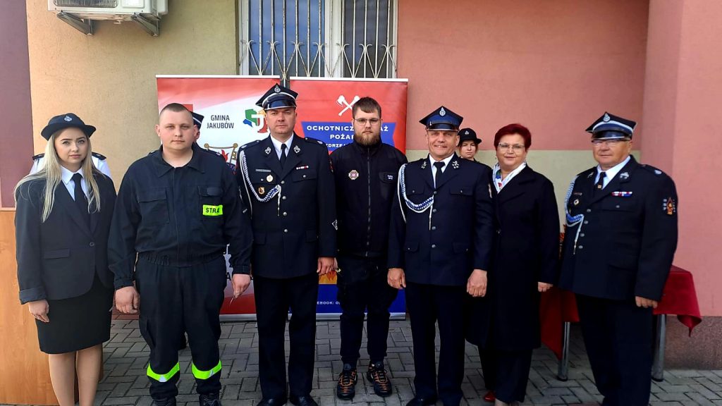 Na zdjęciu grupa osób w mundurach strażackich i formalnych stoi na tle czerwonego banera. Widoczny jest jasnobrązowy budynek. Cztery osoby w czapach strażackich po obu stronach.