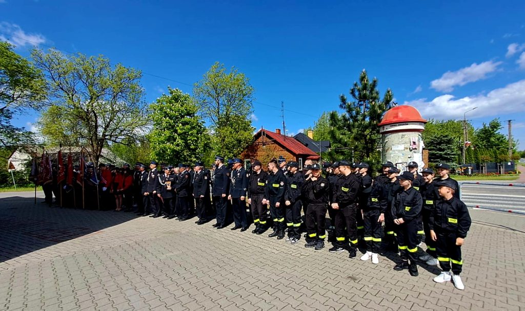 Grupa strażaków w czarnych mundurach stoi na tle zielonych drzew i niebieskiego nieba. W tle widoczny jest czerwony dach budynku. Przed nimi stoją osoby z flagami.