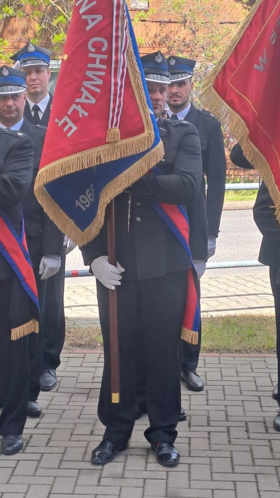 Grupa strażaków w mundurach trzyma czerwone sztandary z frędzlami. W tle drzewa i budynek. Atmosfera uroczysta.