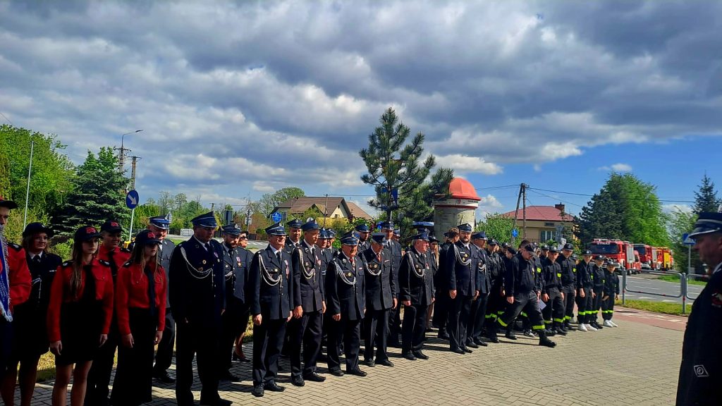 Grupa strażaków w paradnych mundurach stoi na chodniku. Tło to zielone drzewa i niebo z chmurami. W oddali widać budynek i wozy strażackie.