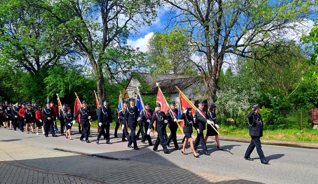 Parada strażaków w mundurach na ulicy z flagami, w tle zielone drzewa i niebieskie niebo z chmurami.