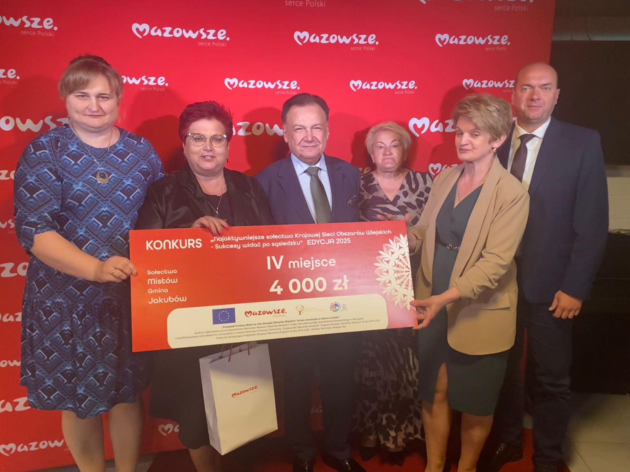 Na czerwonym tle z logo Mazowsze stoi grupa sześciu osób. Trzymają duży czek na kwotę 4000 zł. Osoby są elegancko ubrane, a czek jest głównie czerwony z białym napisem.