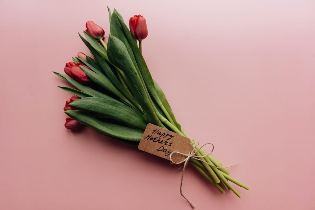 Bukiet czerwonych tulipanów z zielonymi liśćmi na różowym tle. Dołączona brązowa kartka z napisem Happy Mother's Day.