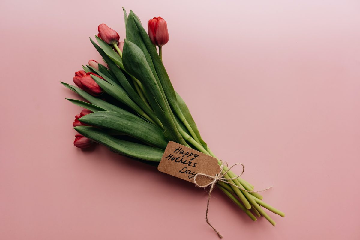 Bukiet czerwonych tulipanów z zielonymi liśćmi na różowym tle. Dołączona brązowa kartka z napisem Happy Mother's Day.