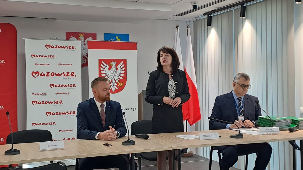 Osoby są przy stole konferencyjnym z mikrofonami. Kobieta stoi, mężczyzna siedzi, drugi mężczyzna czyta dokumenty. W tle polska flaga i logo Mazowsza.