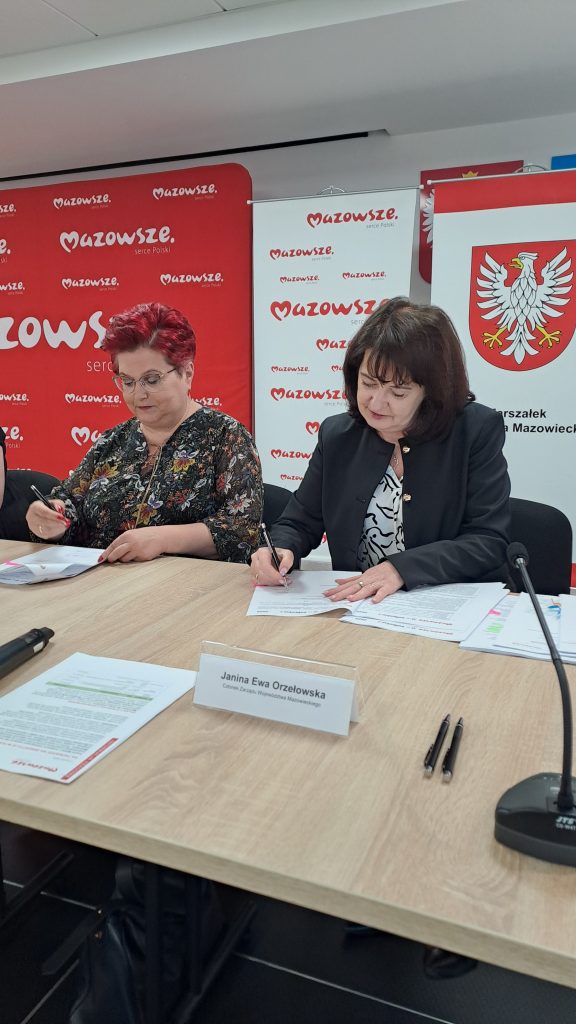 Dwie kobiety podpisują dokumenty przy stole. Tło czerwono-białe z logo Mazowsze oraz herbem. Ubrania ciemne i kolorowe.