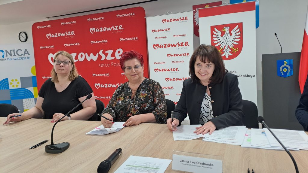 Trzy kobiety siedzą przy stole, podpisując dokumenty. Tło jest czerwone z logo „Mazowsze”. Kobiety mają uśmiechnięte twarze.