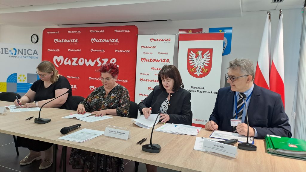 Cztery osoby podpisują dokumenty przy stole. Tło z czerwonym logo Mazowsza. Flagi Polski stoją po prawej stronie.