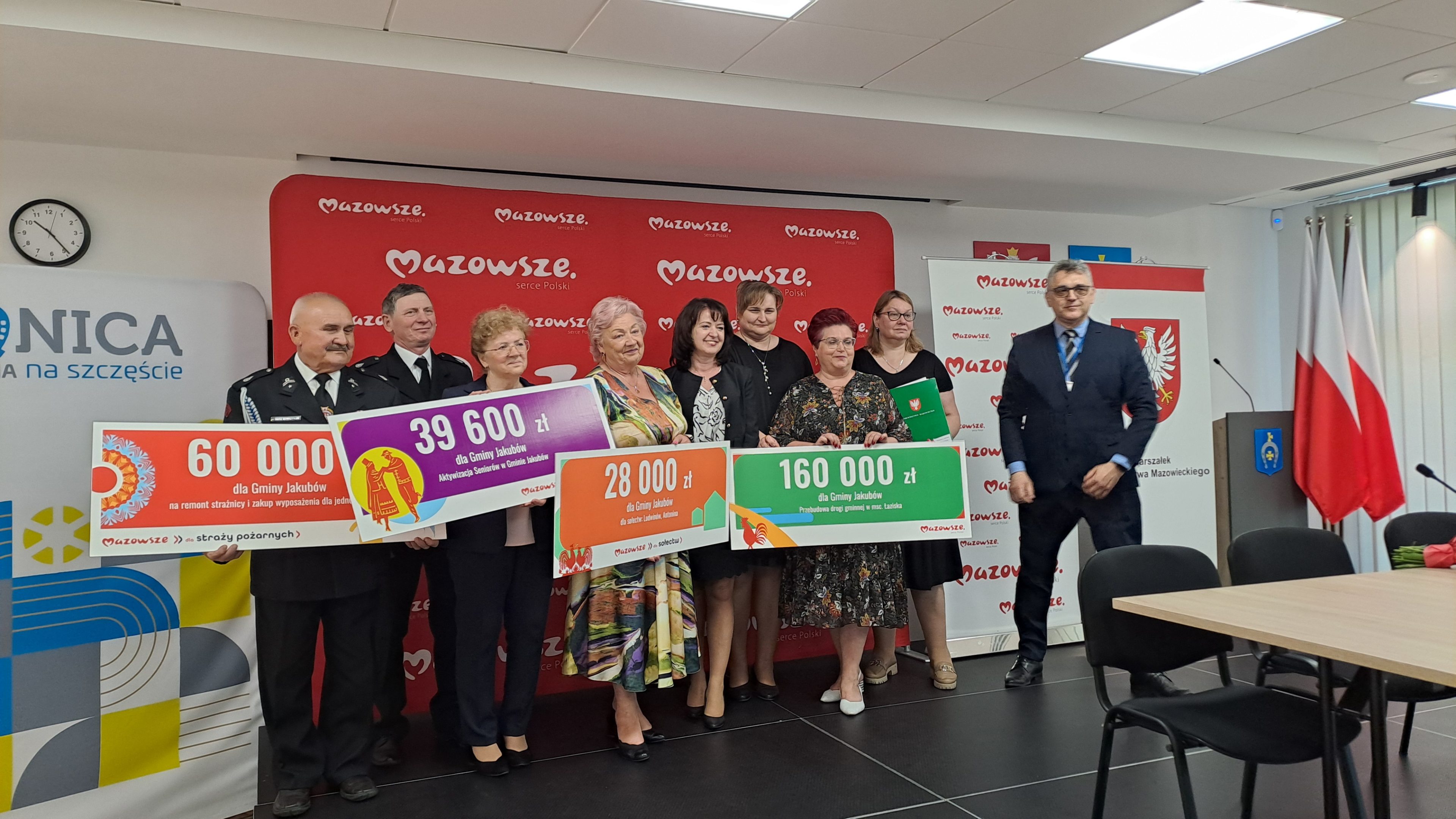 Na zdjęciu grupa osób trzymających kolorowe czeki z kwotami pieniędzy, stojąca na tle czerwonego banera Mazowsze. W tle znajdują się flagi Polski i logo. Dominujące kolory to czerwony, zielony i fioletowy.