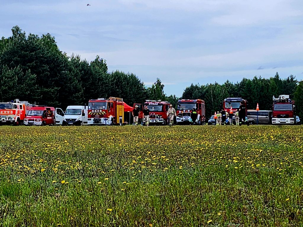 Na łące stoją czerwone strażackie wozy otoczone drzewami. Tło stanowi las, a przed nimi rośnie zielona trawa z żółtymi kwiatami. Widać też ludzi w uniformach strażackich.