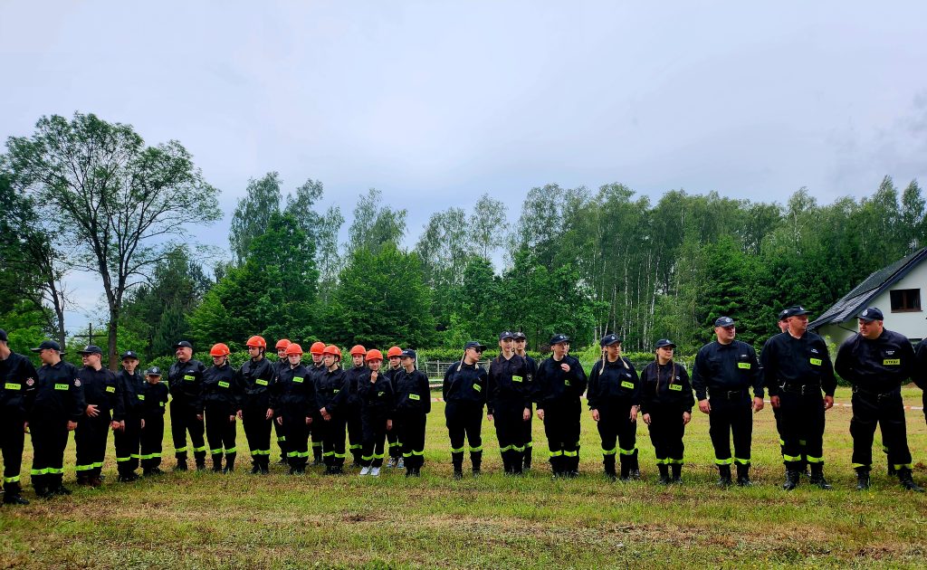 Grupa osób w czarnych uniformach stoi na trawie. Część z nich ma pomarańczowe kaski. W tle widać drzewa i dom.