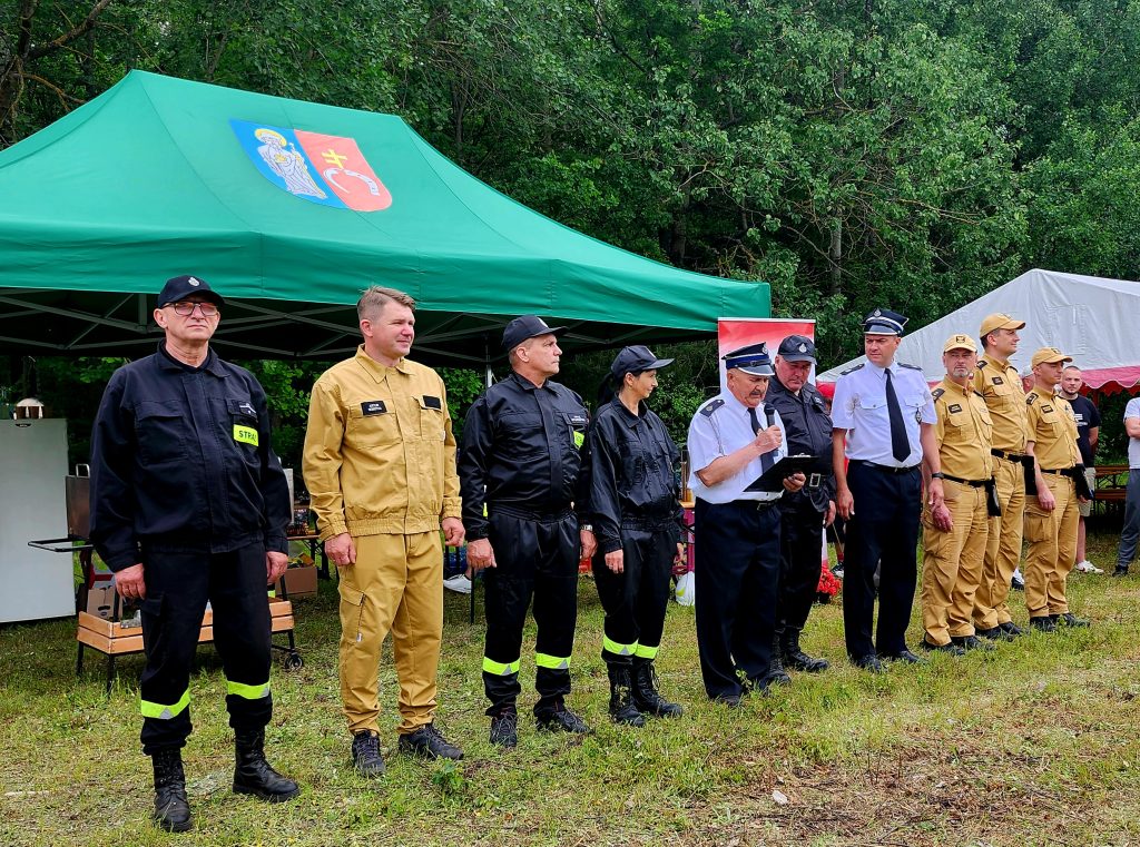 Grupa strażaków stoi przed zielonym namiotem. Mundury strażaków są czarne i żółte. W tle widoczne są drzewa.