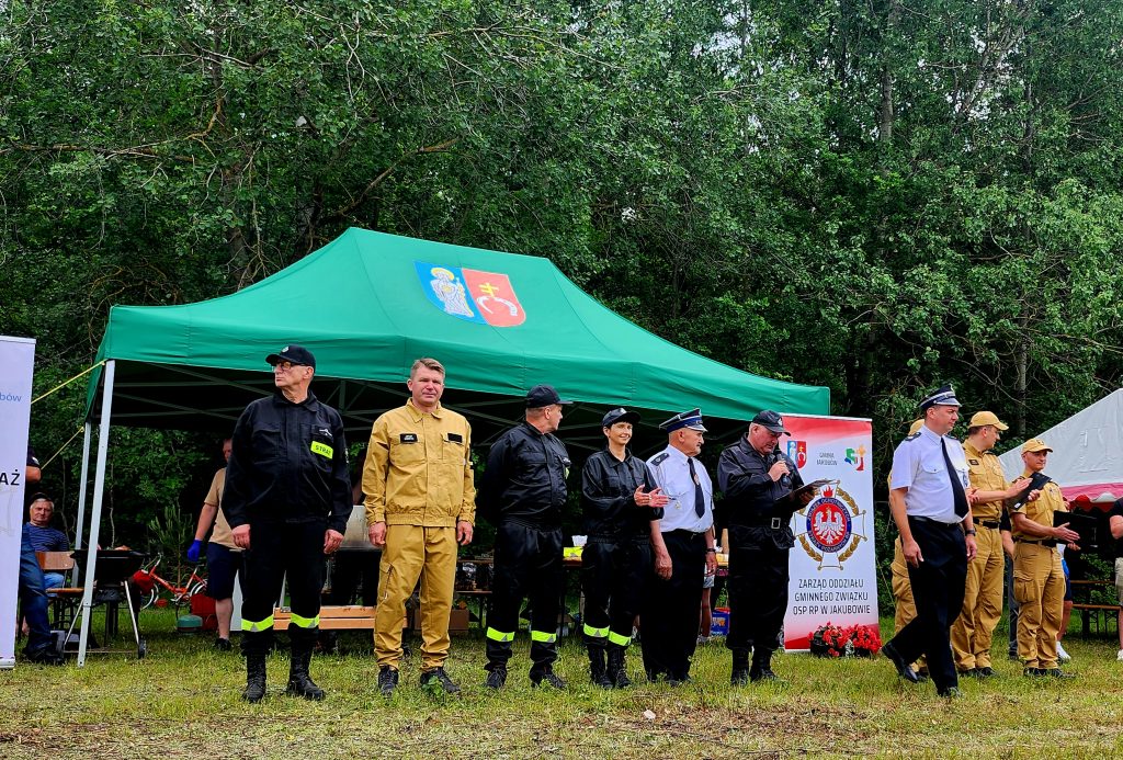 Grupa osób w mundurach stoi przed zielonym namiotem. W tle gęste, zielone drzewa. Banery z napisami i logo.