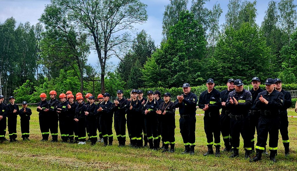 Grupa strażaków w czarnych uniformach stoi na trawie, część w pomarańczowych kaskach. W tle drzewa, niebo zachmurzone. Ludzie klaszczą.