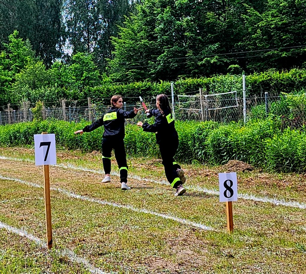 Dwie osoby w czarnych uniformach biegną na trawie, przekazując sobie pałeczkę. Na trawie numery 7 i 8. W tle zielone krzewy i drzewa.