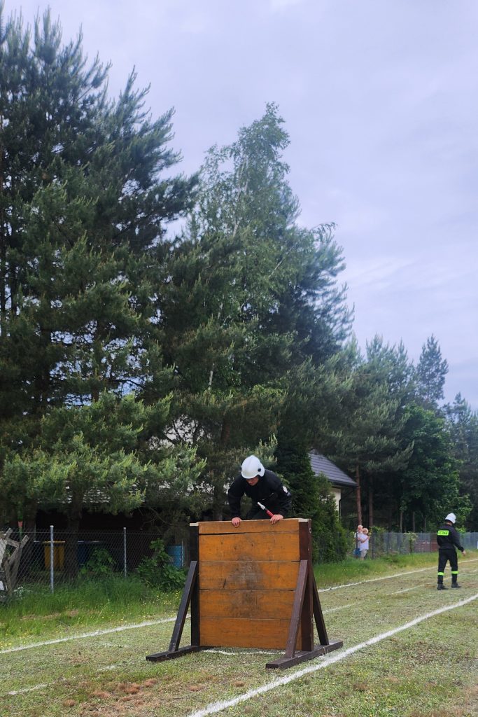 Strażak w czarnym mundurze i białym kasku przeskakuje przez drewnianą przeszkodę. Tło to zielone drzewa i trawnik. Obok stoi inny strażak.
