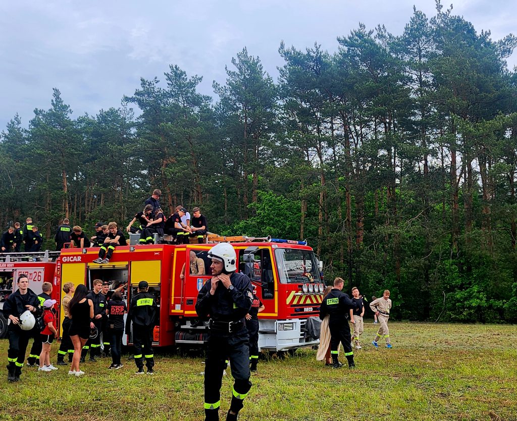 Ludzie w strojach strażackich stoją przy czerwonym wozie strażackim na tle zielonego lasu. Niebo jest pochmurne. Grupa osób siedzi na dachu pojazdu.