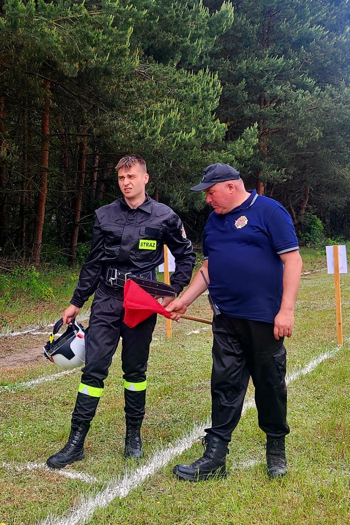 Dwóch strażaków na trawiastej polanie, jeden w czarnym uniformie, drugi w niebieskim. Jeden trzyma hełm, drugi czerwoną flagę. W tle zielony las.