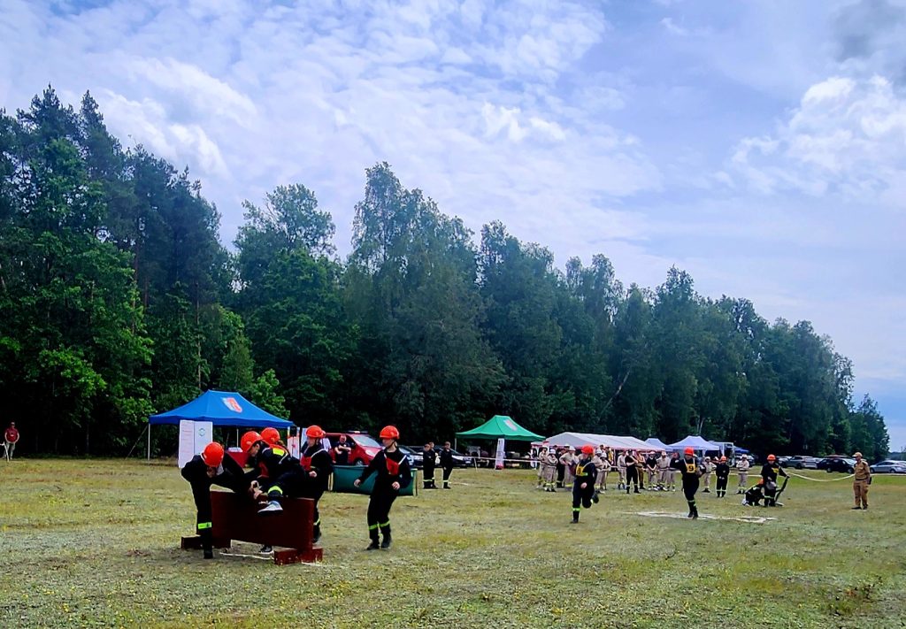 Grupa strażaków w czerwonych kaskach wykonuje ćwiczenia na polanie, w tle las i niebieskie niebo z chmurami. Na polanie widać niebieski i zielony namiot oraz widzów. Tło stanowi zielony las i kilka zaparkowanych samochodów.