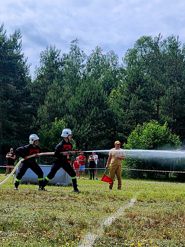 Na zdjęciu dwaj strażacy w czarnych mundurach i białych kaskach trzymają węża, z którego leci woda. W tle las i widownia. Niebo jest pochmurne.