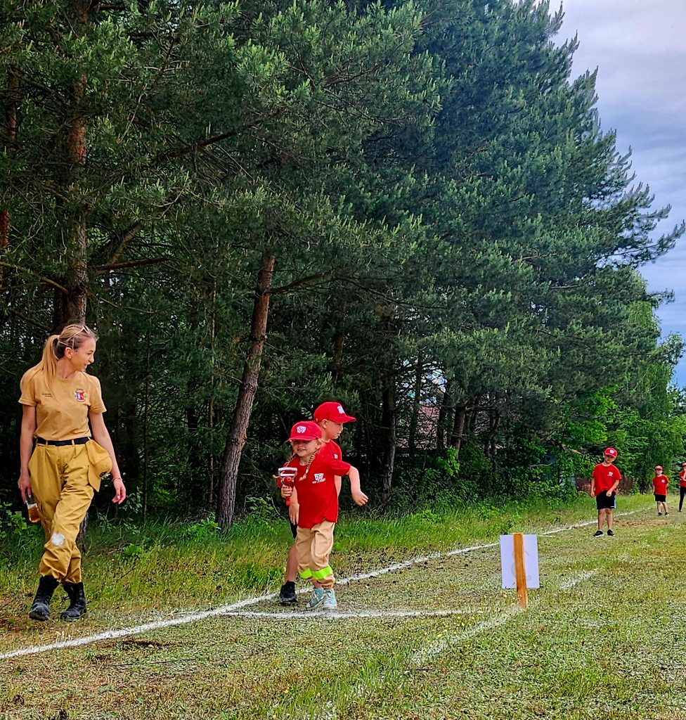 Dwójka dzieci i kobieta w mundurach strażackich idą razem po trawiastym torze. Dzieci są ubrane na czerwono i noszą czapki. Rząd drzew tworzy zielone tło.