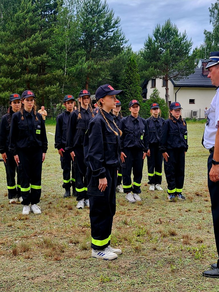 Grupa młodych osób w czarnych mundurach stoi na trawie, słuchając instruktora. W tle zielone drzewa i dom. Niebo pochmurne.