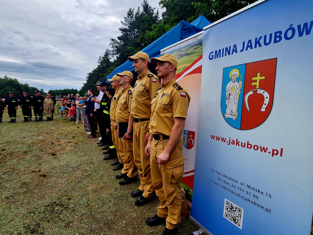 Na zdjęciu widoczni są strażacy w żółtych mundurach stojący na tle niebieskiego namiotu. Obok nich znajduje się baner z napisem Gmina Jakubów i herbem. W tle widać zieleń drzew i pochmurne niebo.