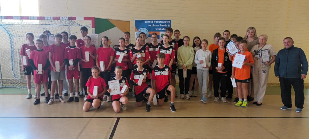Grupa dzieci w kolorowych, sportowych strojach pozuje z nagrodami. W tle znajduje się bramka do piłki ręcznej. Jasne oświetlenie sali gimnastycznej.