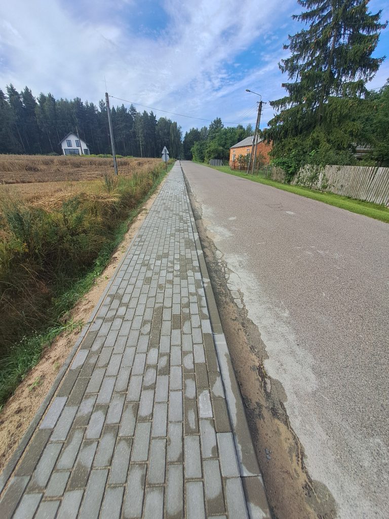 Ścieżka z szarych kostek wzdłuż drogi, otoczona polem i lasem. Na horyzoncie białe i czerwone budynki. Błękitne niebo z chmurami.