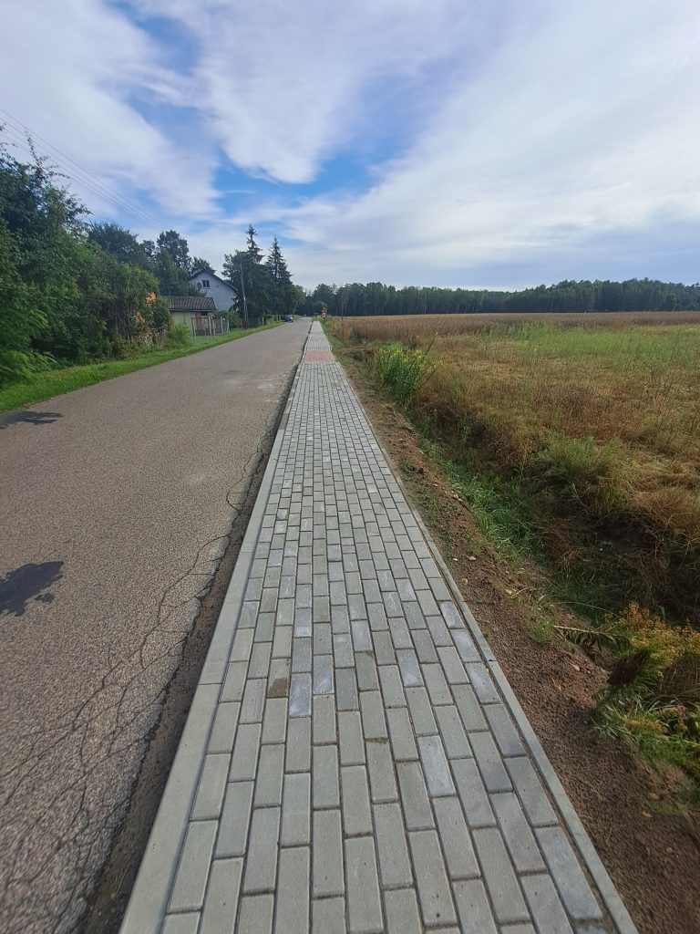 Nowy chodnik z szarej kostki biegnie wzdłuż asfaltowej drogi. Po lewej stronie zielone drzewa i dom, po prawej pole. Niebo pokryte jasnymi chmurami.