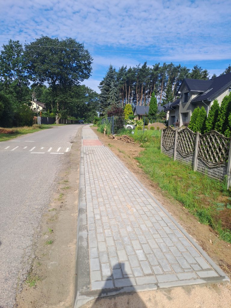 Nowy szary chodnik obok wiejskiej drogi, w tle domy i drzewa. Błękitne niebo z lekkimi chmurami. Zielone trawniki po obu stronach.