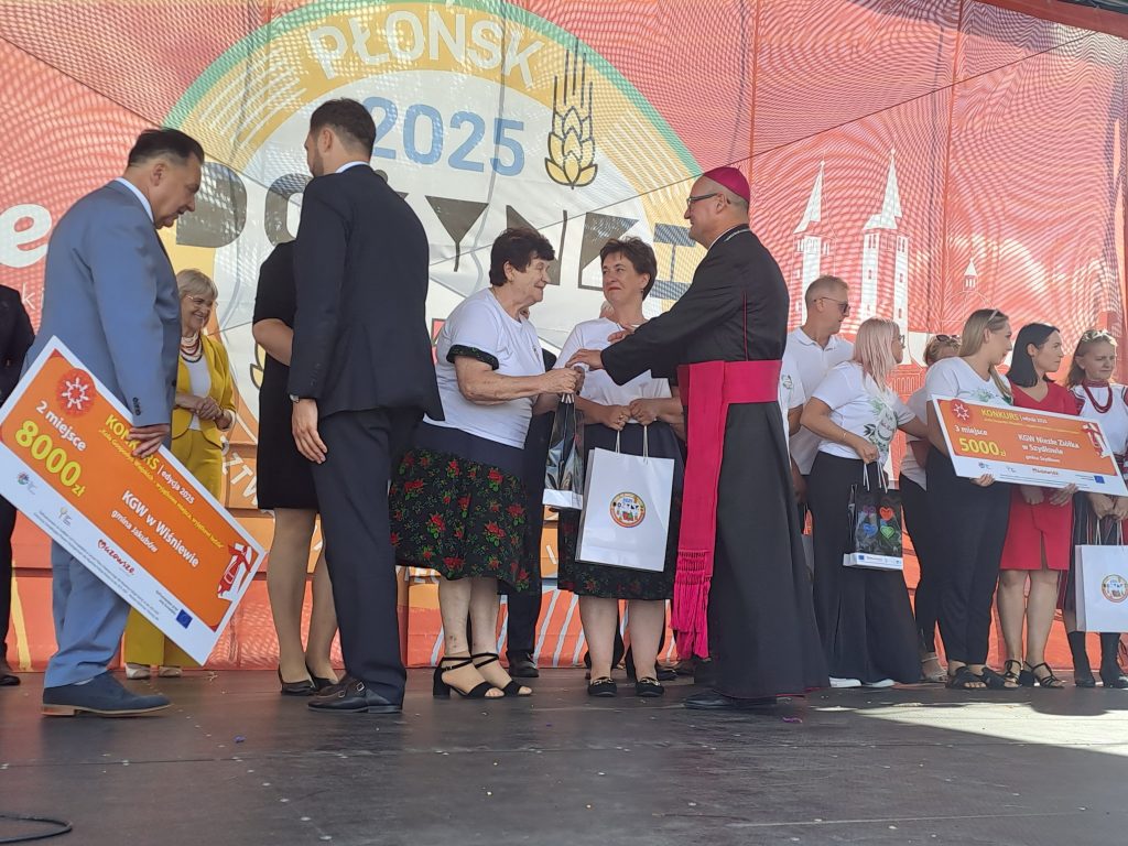 Ludzie na scenie odbierają nagrody z dużymi czekami. Tło jest czerwono-pomarańczowe z logo wydarzenia. Osoba w czarnym stroju duchownym wręcza torby.