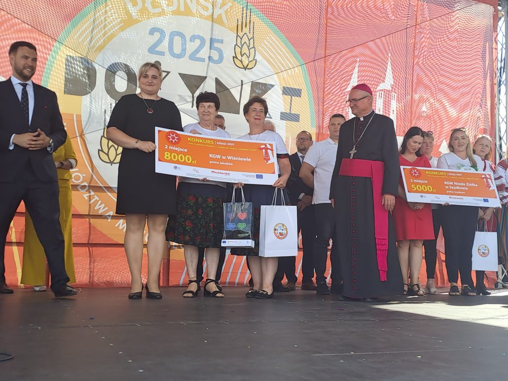 Na scenie grupa ludzi trzyma duże czeki z nagrodami. Tło w kolorze czerwonym z napisem Dożynki 2023. Uczestnicy są elegancko ubrani.