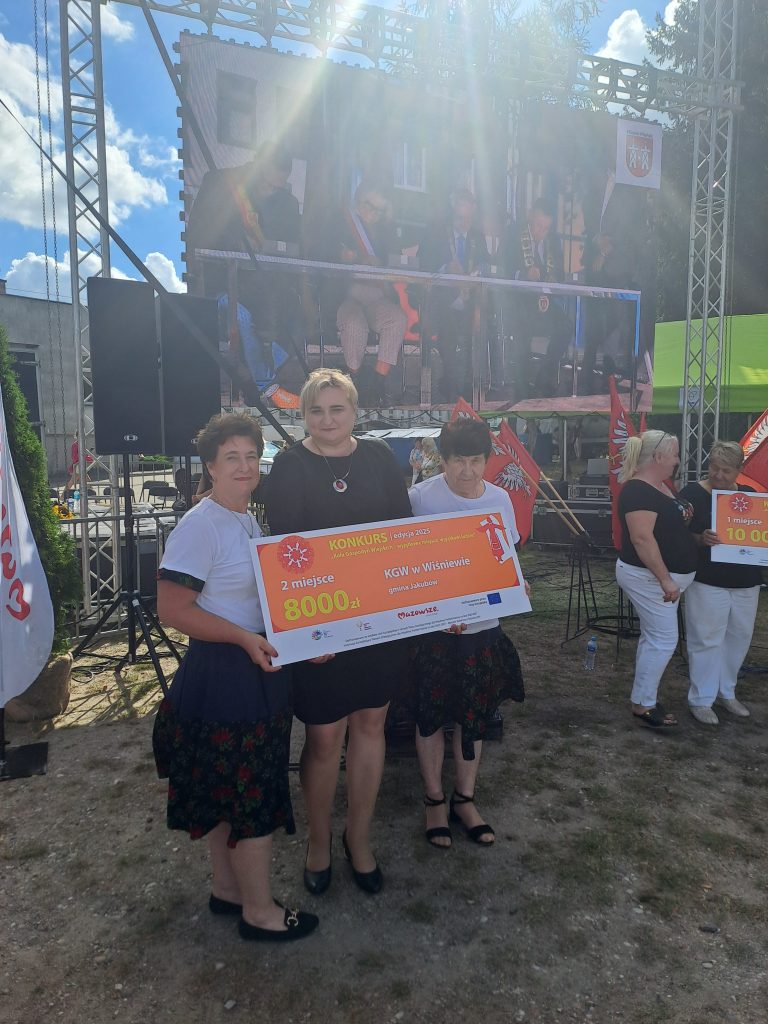 Trzy kobiety trzymają czek na wydarzeniu na świeżym powietrzu. Tło to ekran LED i stalowa konstrukcja. Pogoda jest słoneczna.