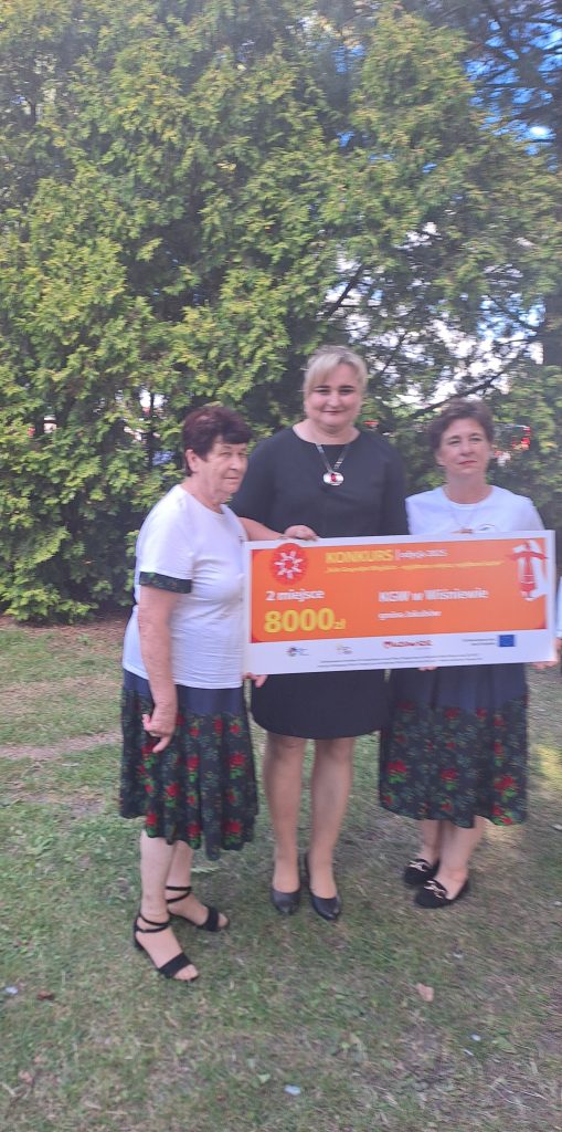Trzy osoby trzymają duży czek na kwotę 8000 zł. Kobiety noszą jednolite stroje ludowe. W tle zielone krzewy.