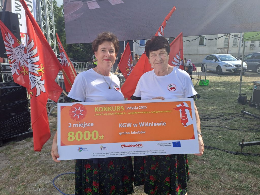 Na zdjęciu dwie uśmiechnięte kobiety trzymają duży, pomarańczowy czek na kwotę 8000 zł. W tle czerwone flagi z białymi orłami i fragment sceny. Obie mają na sobie białe koszulki, a na trawie stoi